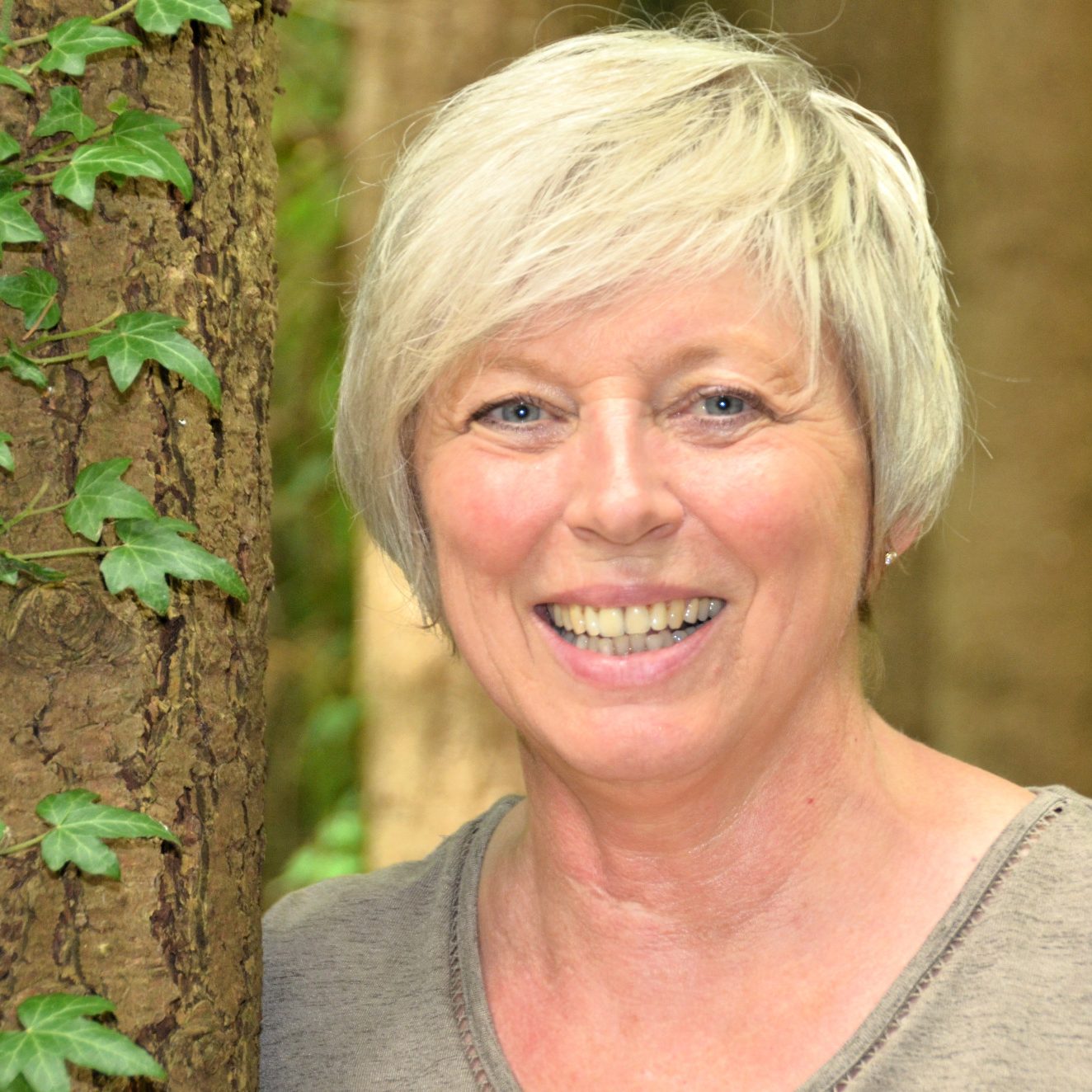 Portrait Bettina Hermey Lächelnde Frau mit kurzer, grauer Frisur, steht vor einem Baum in einem Wald.